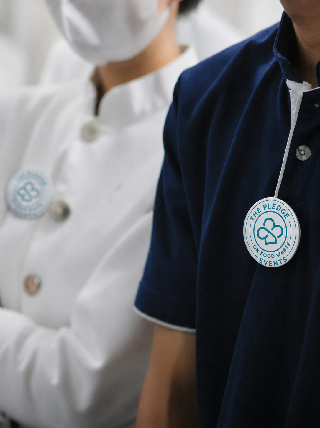 Two hospitality staff members wearing “The PLEDGE on Food Waste Events” badges on their uniforms, one in focus in a dark shirt and one blurred in the background.