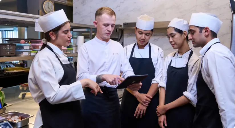 A diverse team of five chefs in uniforms standing in a professional kitchen while looking at and discussing information displayed on a digital tablet held by the head chef.