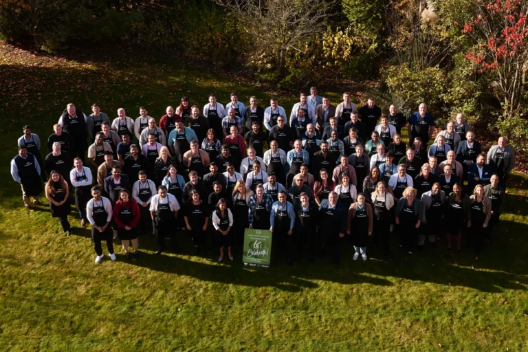 High-angle outdoor group photo of dozens of people wearing black aprons, standing on a green lawn with a "Let's Celebrate" sign in the center.