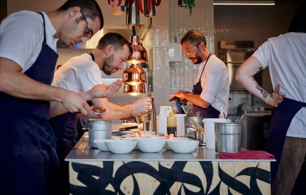 Certified restaurant chefs plating dishes in the open kitchen at Refettorio Geneva. The food waste certification restaurant demonstrates zero waste kitchen practices through collaborative culinary preparation and sustainable gastronomy operations.